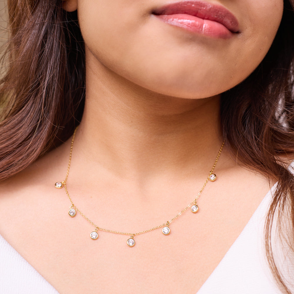 A close-up of a woman wearing a delicate gold necklace with five small clear round gemstones spaced along the chain.