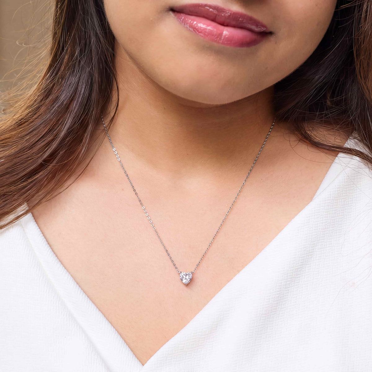 A close-up of a woman wearing a delicate silver chain necklace with a small heart-shaped clear gemstone pendant, paired with a white V-neck top.