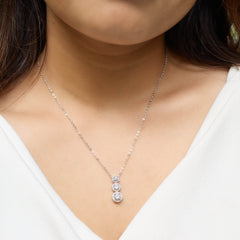 A close-up of a model wearing a delicate silver chain necklace with a pendant featuring three stacked round clear gemstones, set against a white V-neck top.